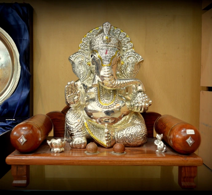 Pure silver Lord Ganesha idol with gold highlights seated on a wooden chowki throne with side bolsters.